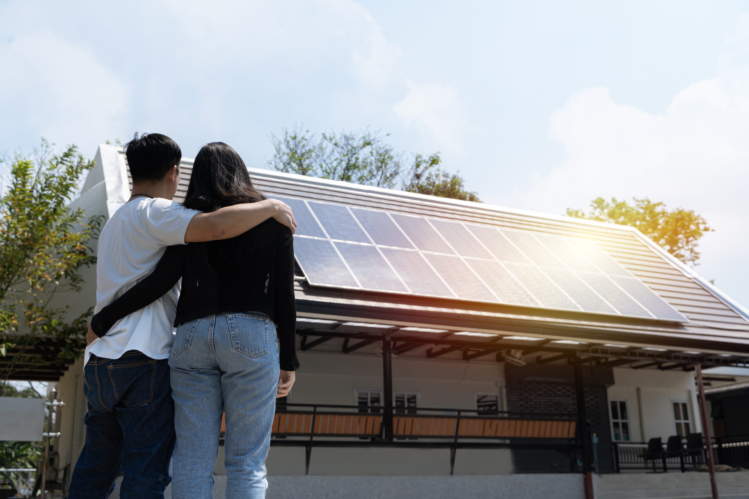 Happy couple standying near their house with solar panels. Alternative energy, saving resources and sustainable lifestyle concept. Renewable energy electricians in Farnham